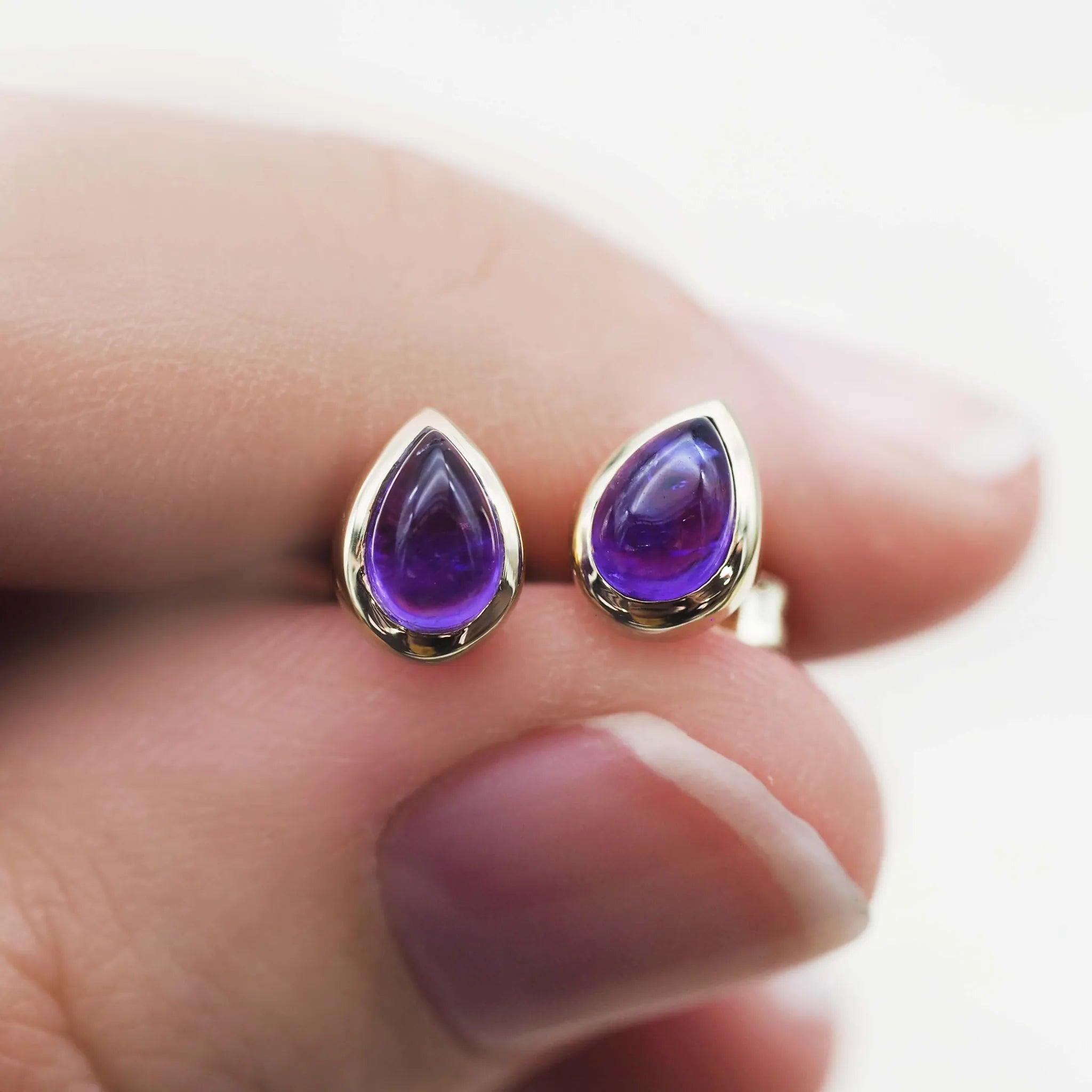 Purple teardrop earrings with gold accents held between fingers against a blurred background