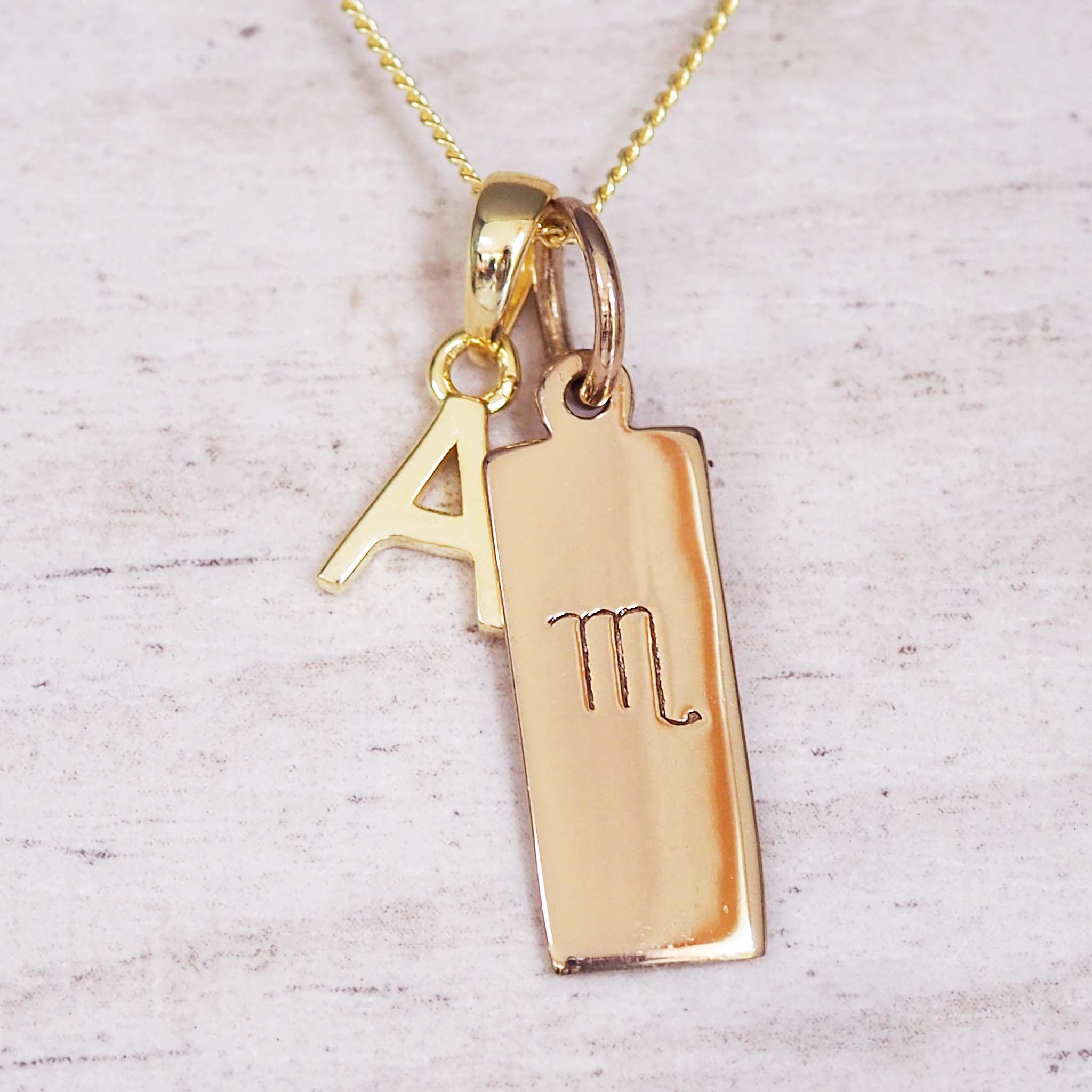 Gold necklace with a rectangular pendant featuring an engraved letter 'A' on a light wooden background