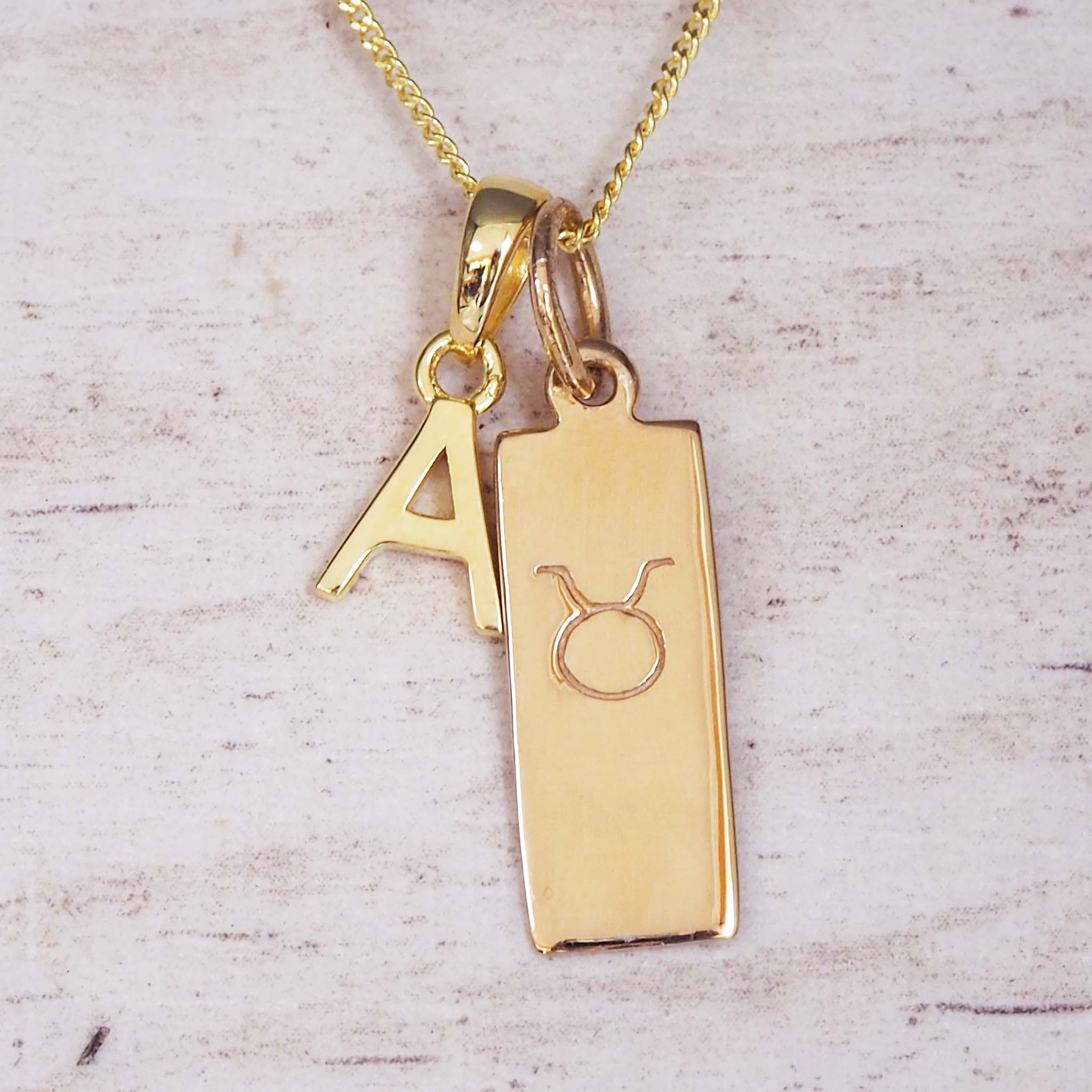 Gold necklace with 'A' and Taurus symbol pendant on a light wooden background