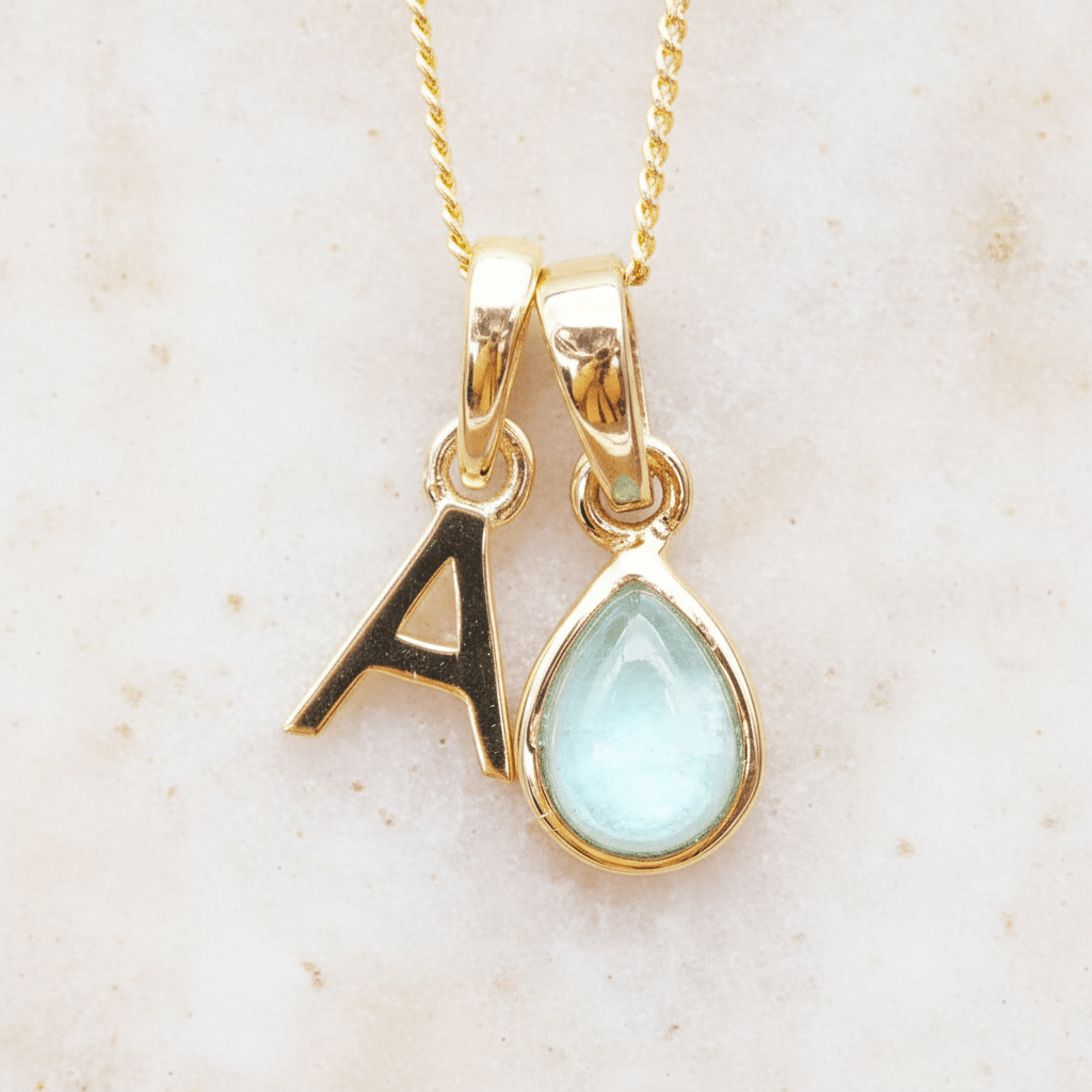Gold necklace with 'A' initial pendant and a march birthstone natural aquamarine gemstone pendant on a beige background