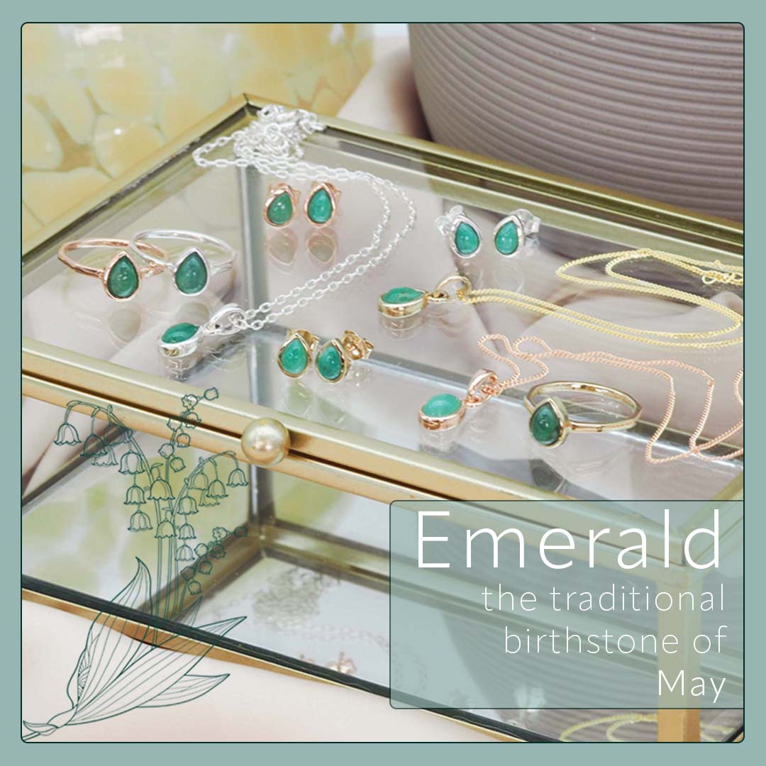 Jewelry set with emerald stones on a glass surface, featuring text about emeralds as the traditional birthstone of May.