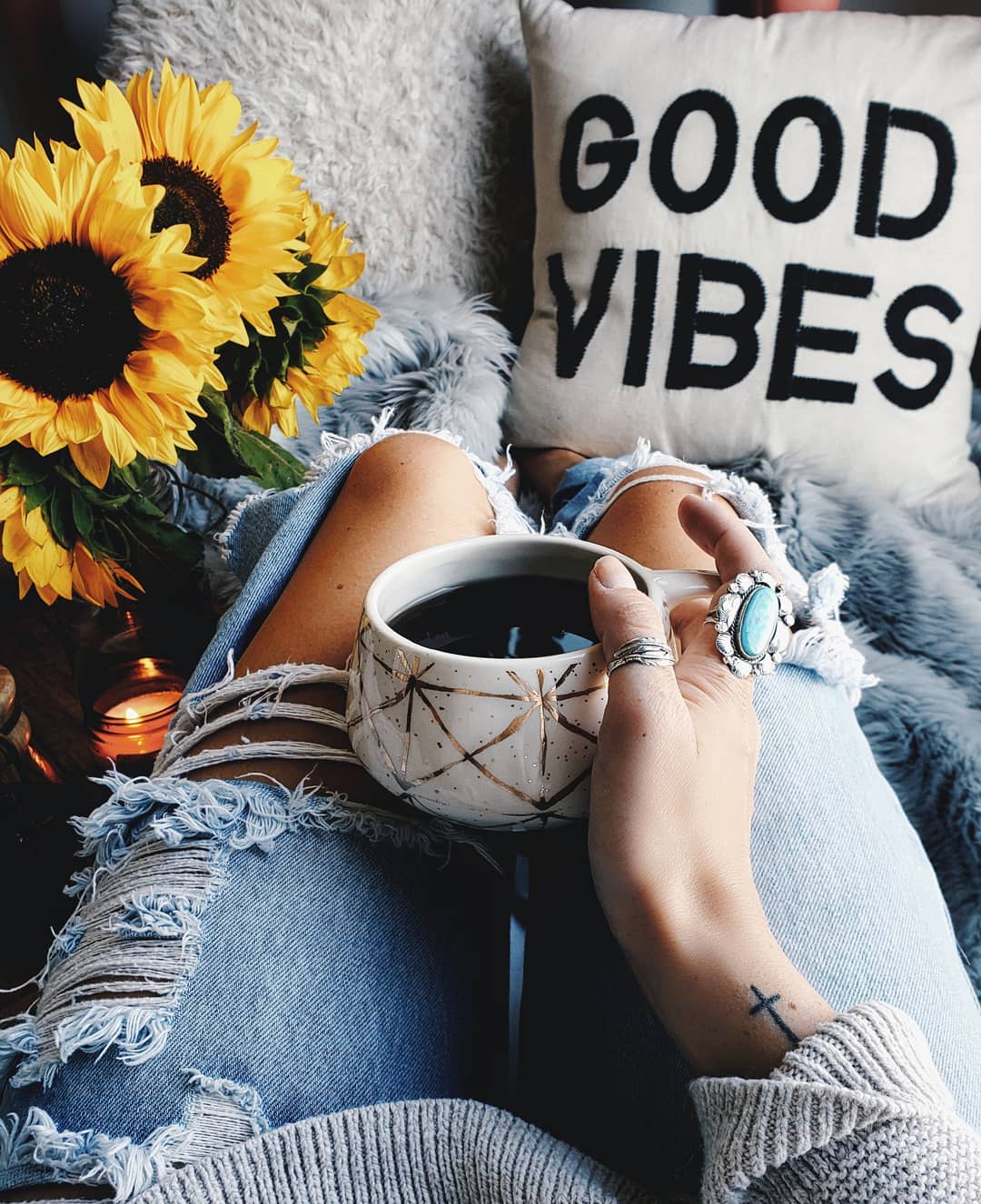 Person holding a mug, wearing a silver turquoise ring, with sunflowers and a 'Good Vibes' pillow in the background