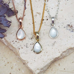 Three teardrop-shaped pendants on gold, rose gold, and silver chains against a stone background.