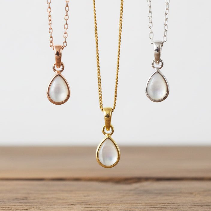 Three teardrop-shaped pendants on gold, rose gold, and silver chains against a stone background.