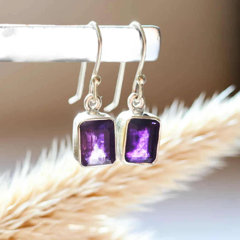 Silver earrings with purple gemstones on a white surface with dried grass.