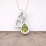 Silver necklace with a 'D' pendant and a teardrop-shaped green gemstone on a wooden surface.