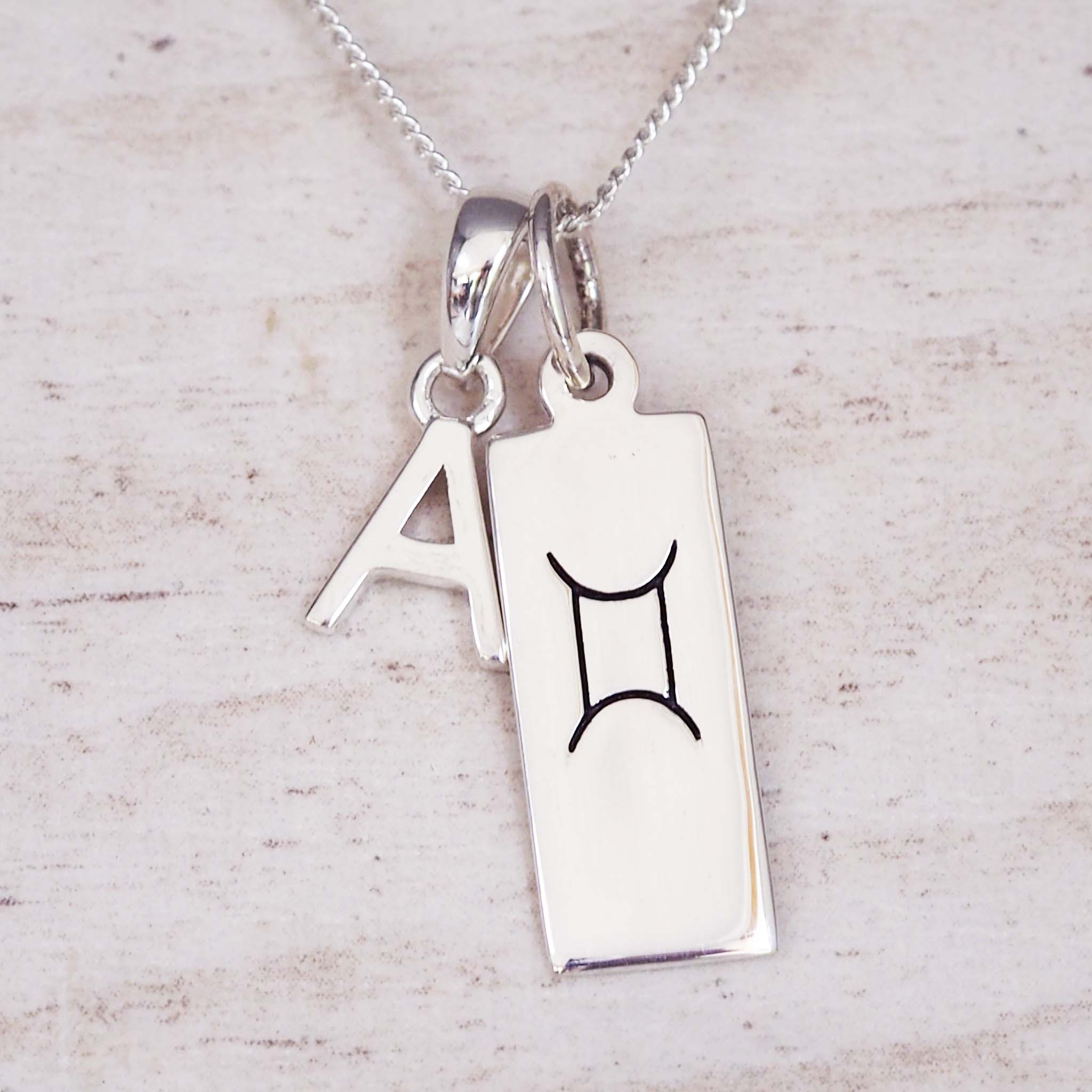Silver necklace with a rectangular pendant featuring a black astrological symbol and a letter 'A'.