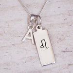 Silver necklace with a rectangular pendant featuring a black symbol and an initial 'A' on a light wooden background.