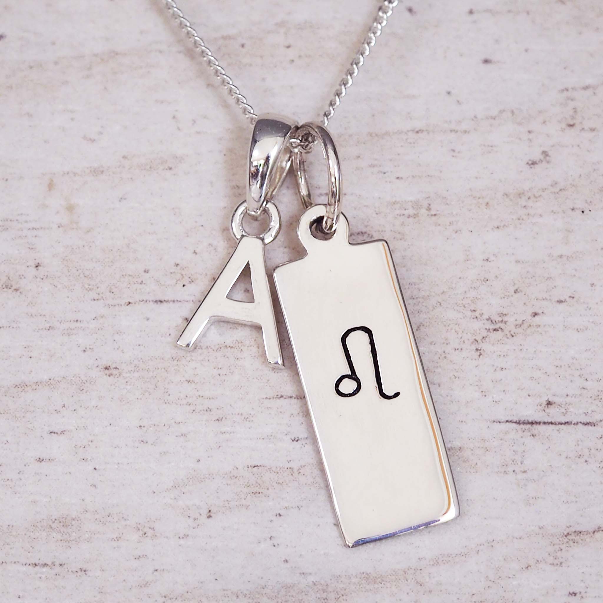 Silver necklace with a rectangular pendant featuring a black symbol and an initial 'A' on a light wooden background.