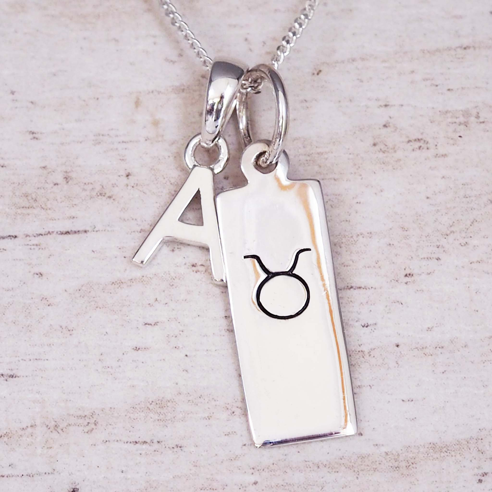 Silver necklace with a rectangular pendant featuring a black symbol and an initial on a light wooden background.