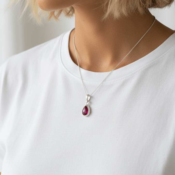 Person wearing a silver necklace with a teardrop-shaped pendant featuring a red gemstone.
