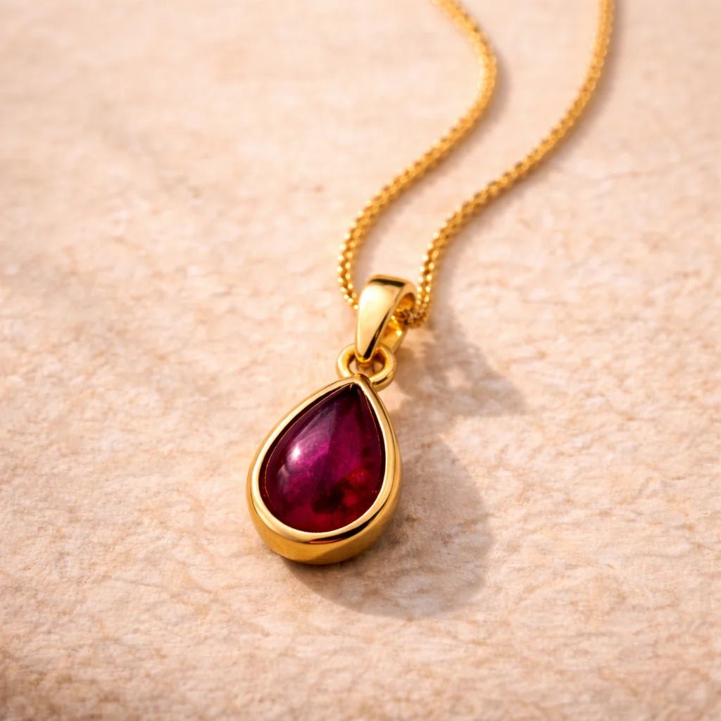 Gold January Birthstone Necklace with a red teardrop natural garnet gemstone pendant on a beige surface