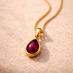 Gold January Birthstone Necklace with a red teardrop natural garnet gemstone pendant on a beige surface