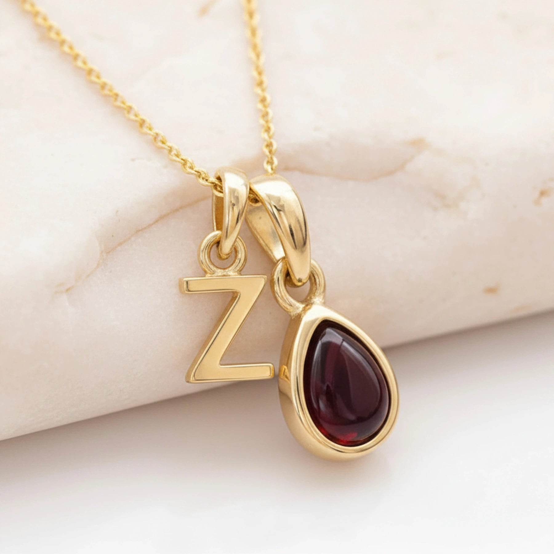 Gold necklace with 'Z' pendant and teardrop-shaped natural garnet gemstone on light stone background