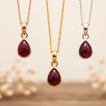 Three January Birthstone Necklaces hanging with teardrop-shaped garnet gemstone pendants on silver, gold and rose gold chains against a beige background