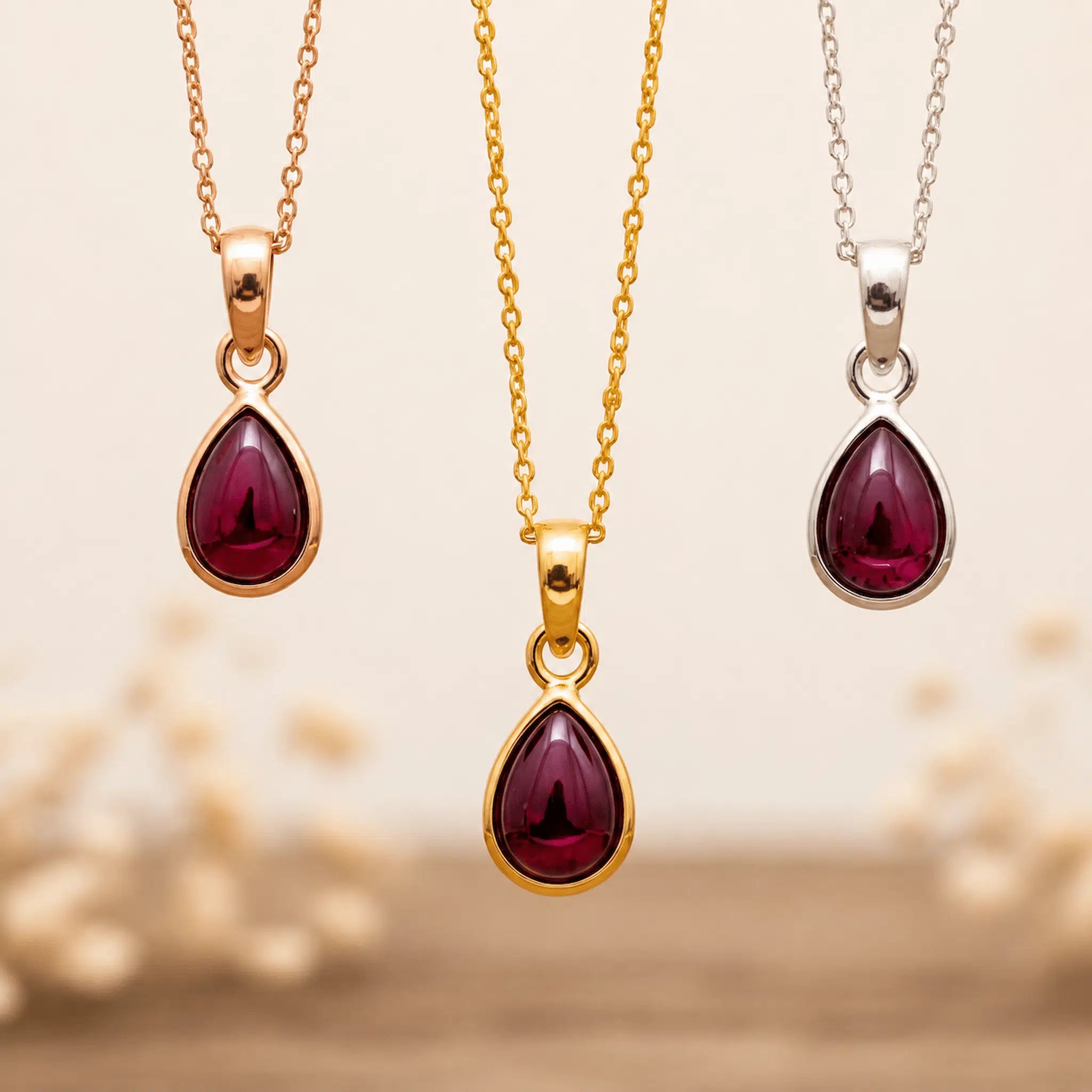 Three January Birthstone Necklaces hanging with teardrop-shaped garnet gemstone pendants on silver, gold and rose gold chains against a beige background