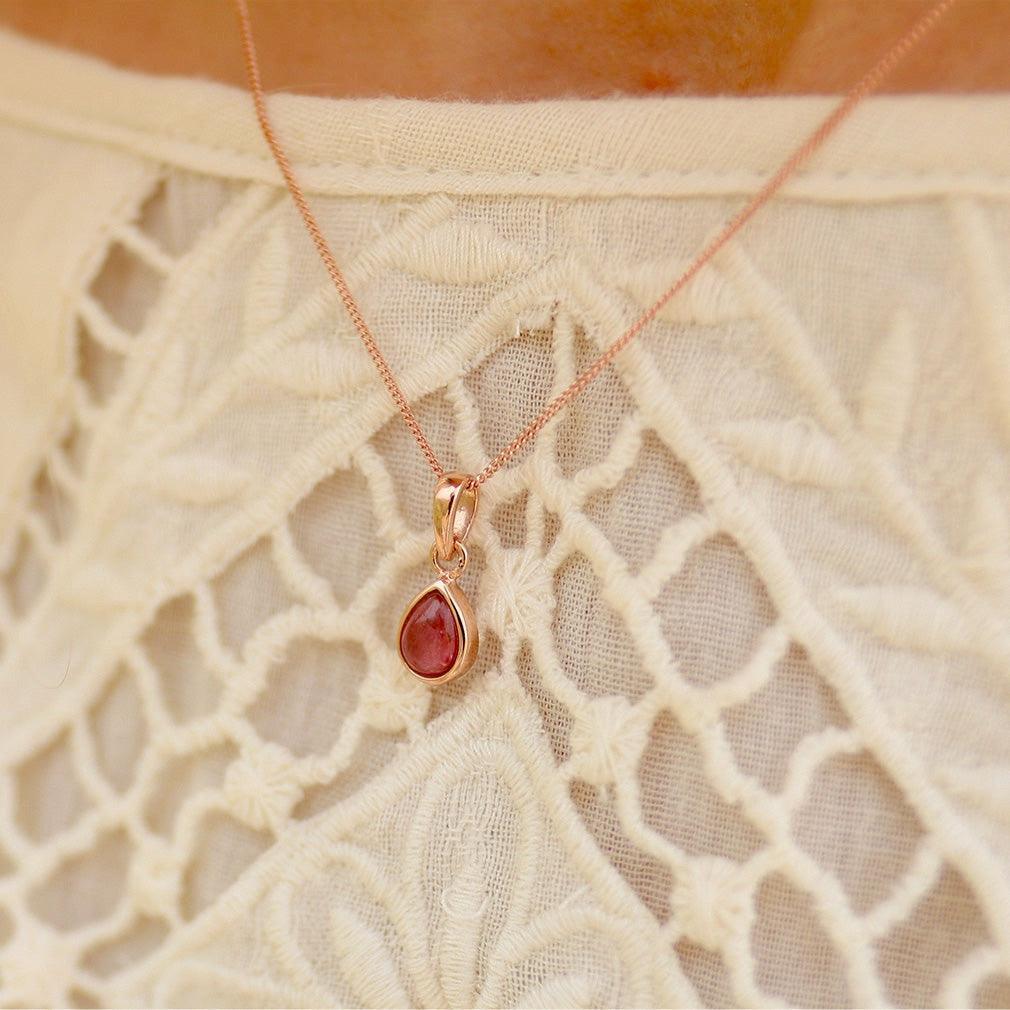 A close-up of a delicate rose gold necklace with a teardrop-shaped red gemstone pendant, worn over a cream-colored lace top.