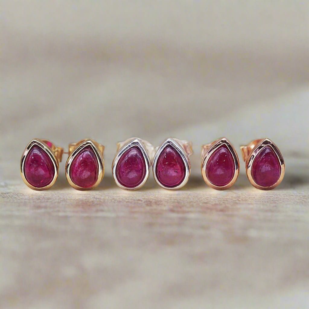Three Pair of tear drop stud earrings in rose fold, silver and gold with red ruby gemstones on a beige background