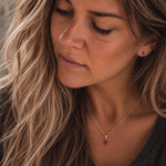 A close-up lifestyle shot of a woman wearing a sterling silver july jewellery set featuring a delicate silver necklace with a teardrop-shaped red natural ruby gemstone pendant and a matching red gemstone stud ruby earring.