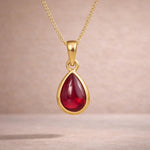 Gold necklace with a red teardrop natural ruby gemstone pendant on a beige background