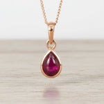 Rose gold necklace with a teardrop-shaped ruby pendant on a light wooden surface.