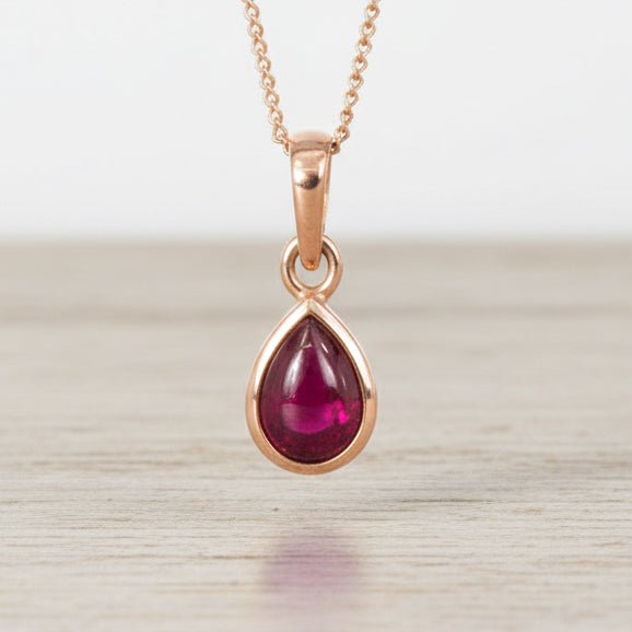 Rose gold necklace with a teardrop-shaped ruby pendant on a light wooden surface.