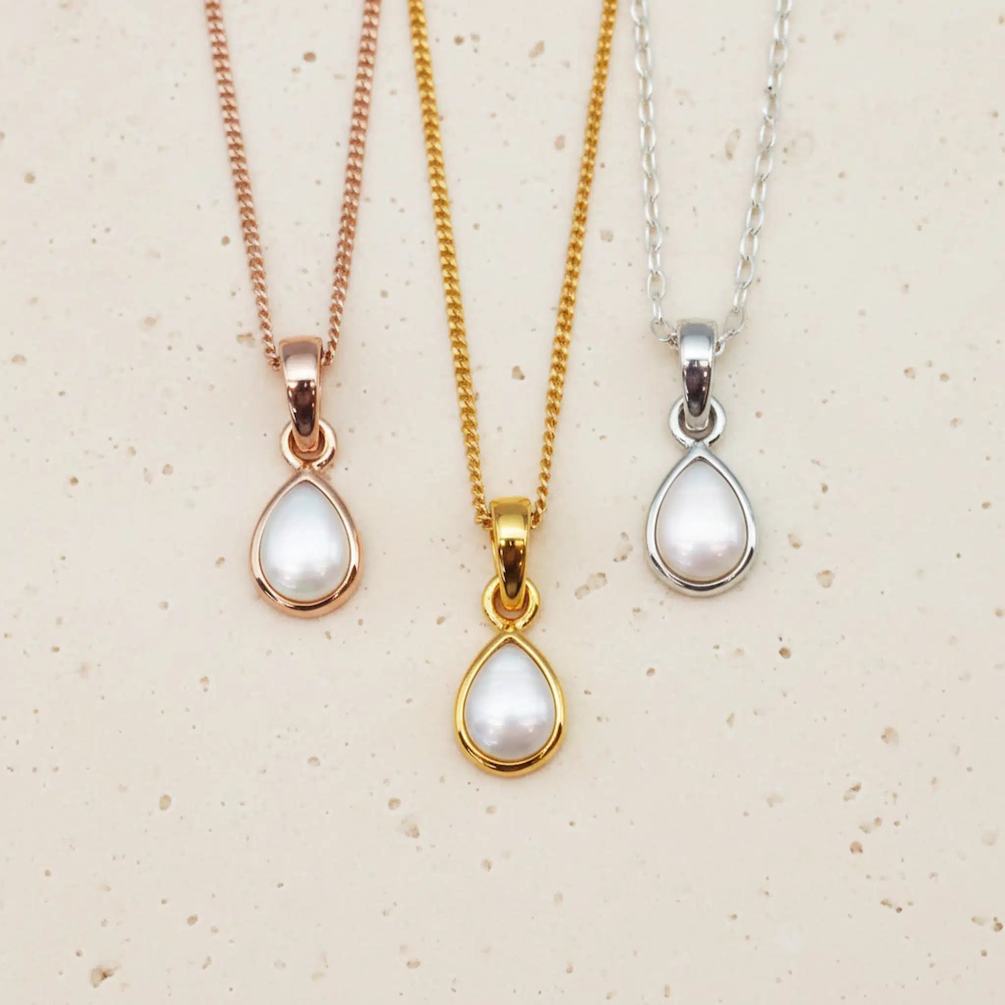 Three pearl pendant necklaces in gold, rose gold, and silver on a beige background