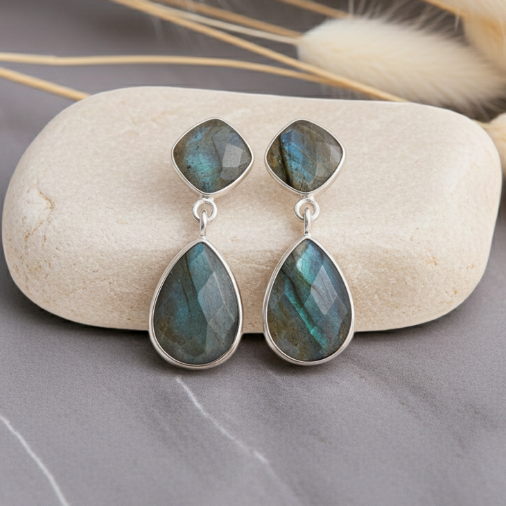 Teardrop-shaped earrings with green gemstones on a stone surface with dried grasses in the background