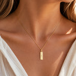 A close-up of a model wearing a delicate gold chain necklace with a small rectangular gold pendant engraved with an astrological leo zodiac symbol, styled with a white top.
