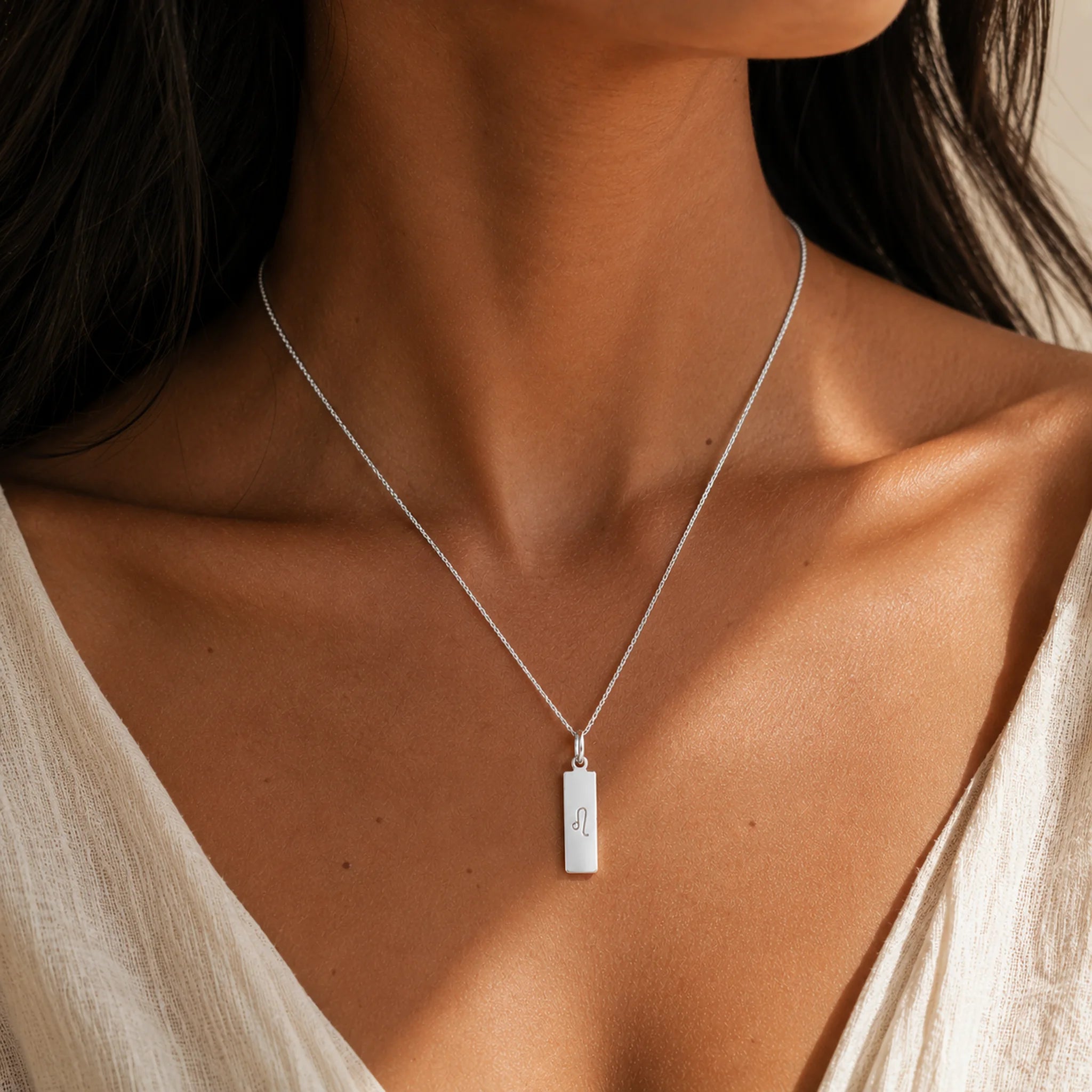 A close-up of a model wearing a delicate silver chain leo necklace with a small rectangular pendant engraved with the leo zodiac line glyph symbol.