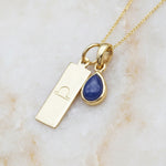 Gold necklace with a blue teardrop sapphire pendant and a rectangular libra zodiac pendant on a warm white stone background.