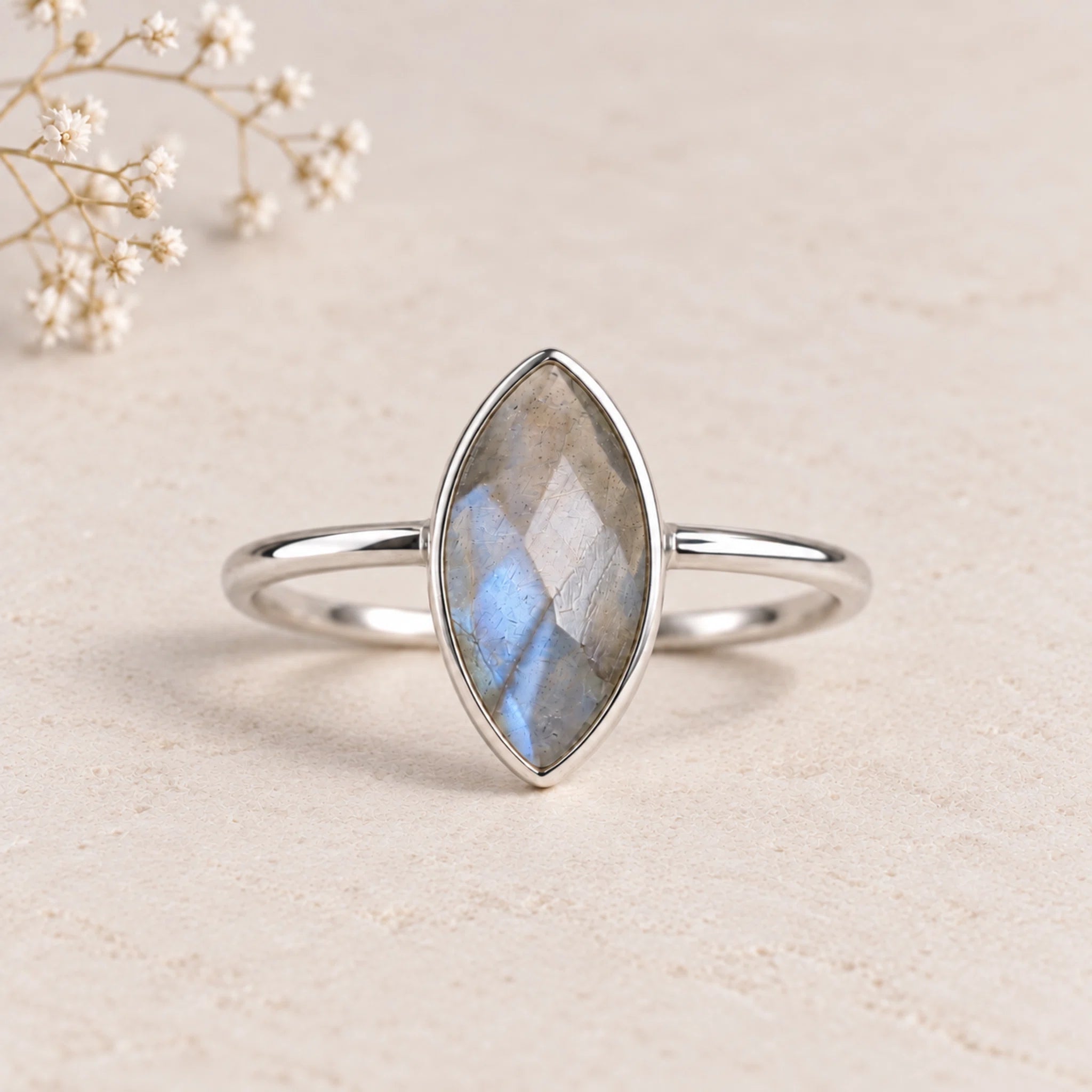 A silver ring with a marquise-cut labradorite gemstone, displayed on a textured beige surface with small white baby's breath flowers in the corner.