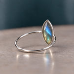 A silver ring with a marquise-cut iridescent labradorite gemstone, resting on a neutral beige surface with a dark teal background.