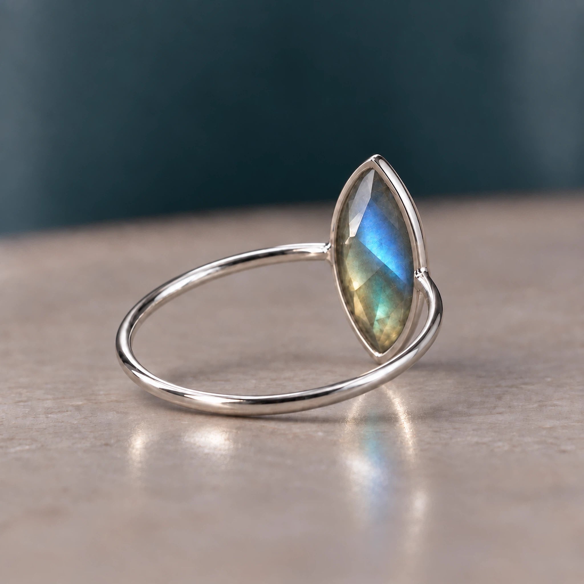 A silver ring with a marquise-cut iridescent labradorite gemstone, resting on a neutral beige surface with a dark teal background.