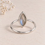 A silver ring with a marquise-cut labradorite gemstone, displayed on a textured beige surface with small dried flowers in the corner, showing open back
