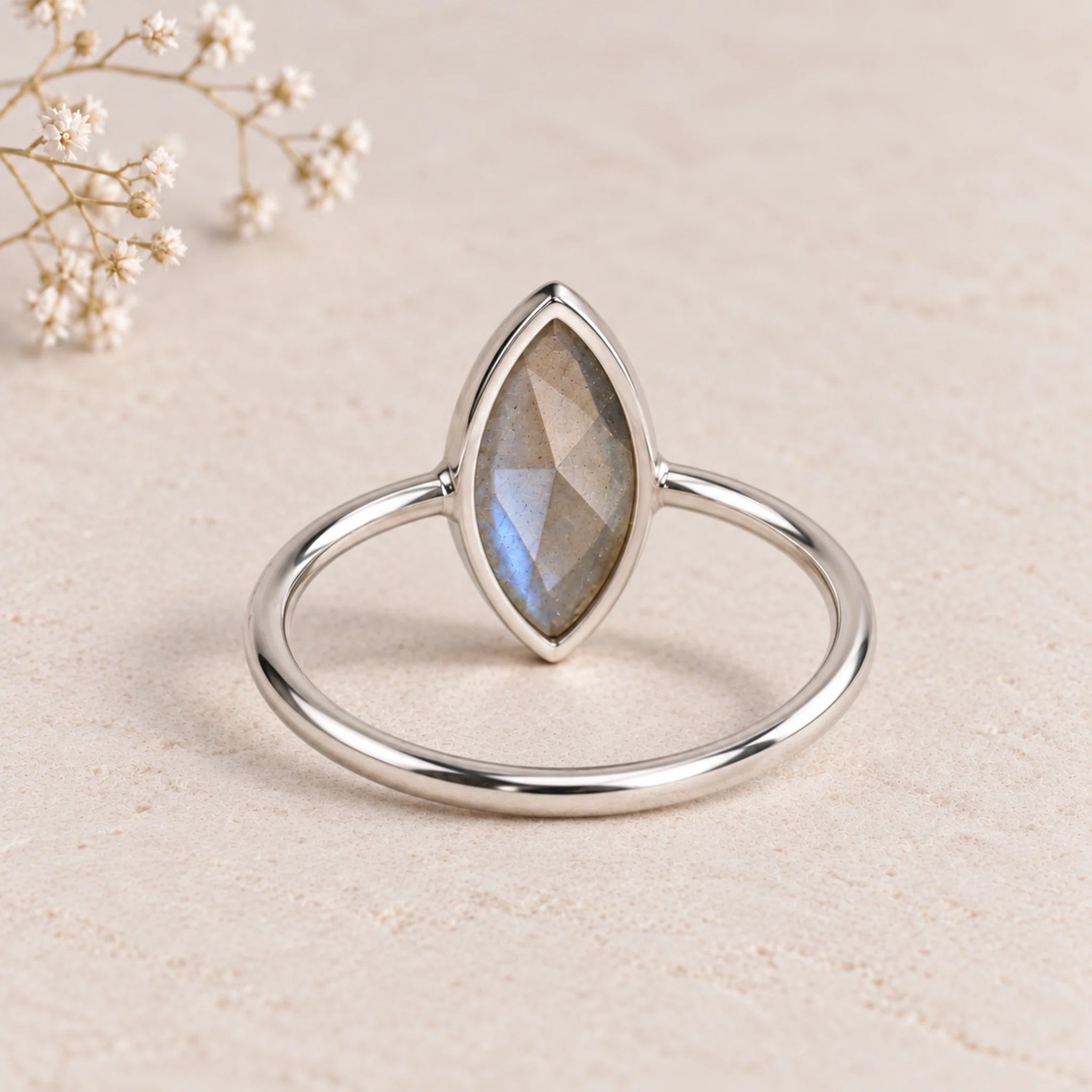 A silver ring with a marquise-cut labradorite gemstone, displayed on a textured beige surface with small dried flowers in the corner, showing open back