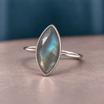 A sterling silver ring featuring a marquise-cut labradorite gemstone with iridescent blue, green, and gray flashes, displayed on a neutral surface with a dark teal background.