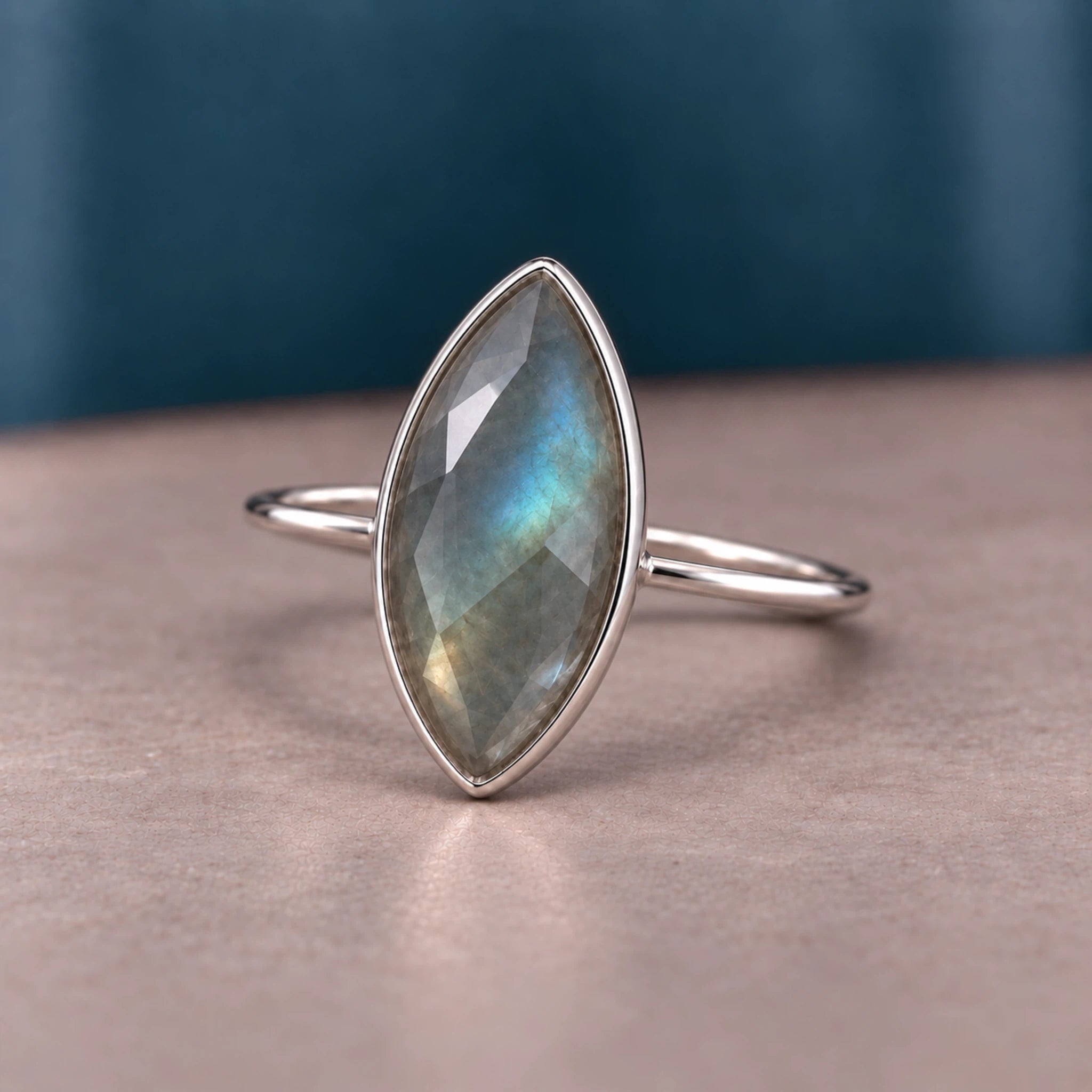 A sterling silver ring featuring a marquise-cut labradorite gemstone with iridescent blue, green, and gray flashes, displayed on a neutral surface with a dark teal background.