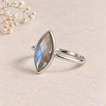 A silver ring featuring a marquise-cut labradorite gemstone with iridescent blue, gray, and gold flashes, displayed on a textured beige surface with small white baby's breath flowers in the corner.