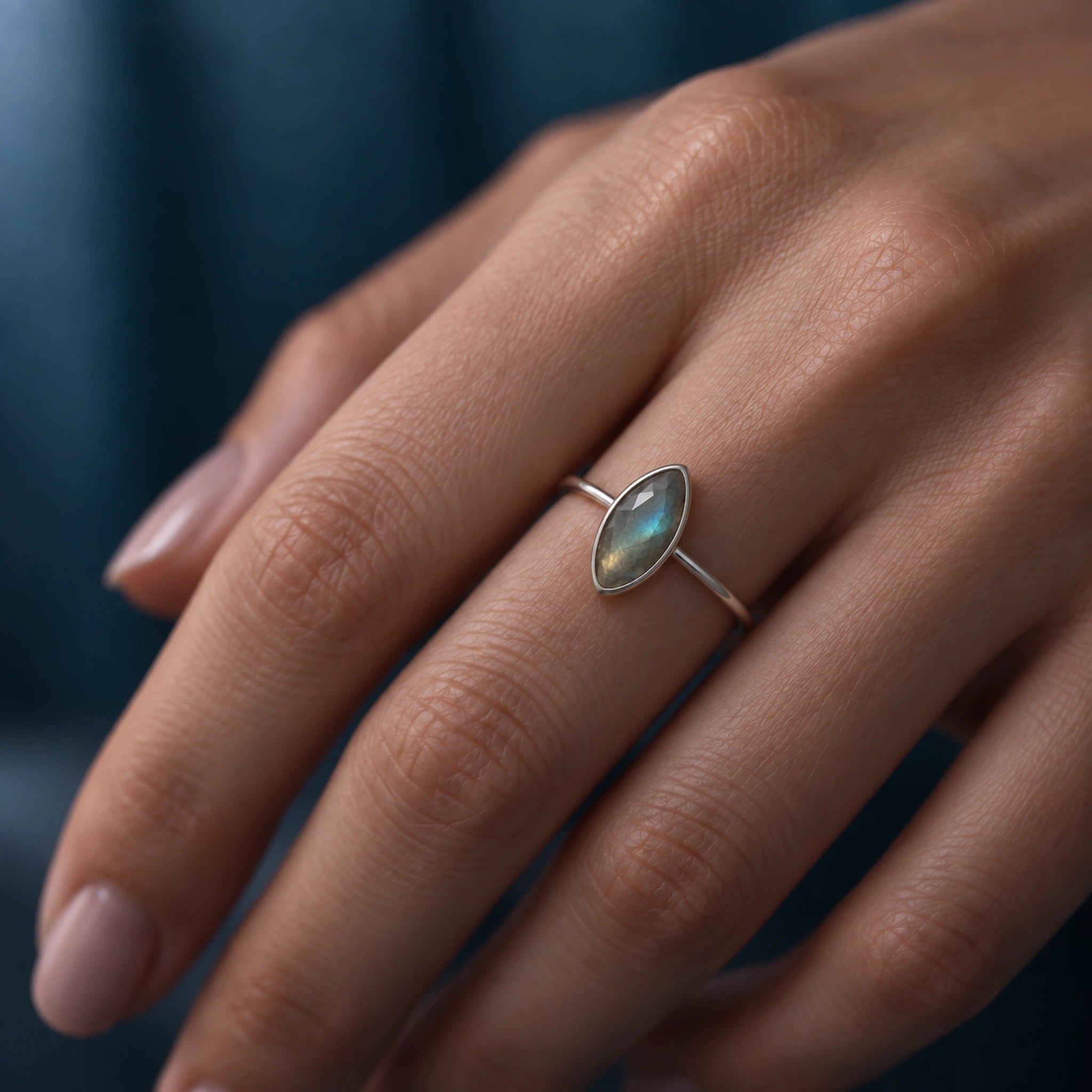 A close-up of a hand wearing a silver ring with a marquise-cut iridescent stone, set against a dark blue background.