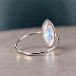A silver ring featuring a marquise-cut iridescent moonstone gemstone, resting on a neutral beige surface with a dark teal background.