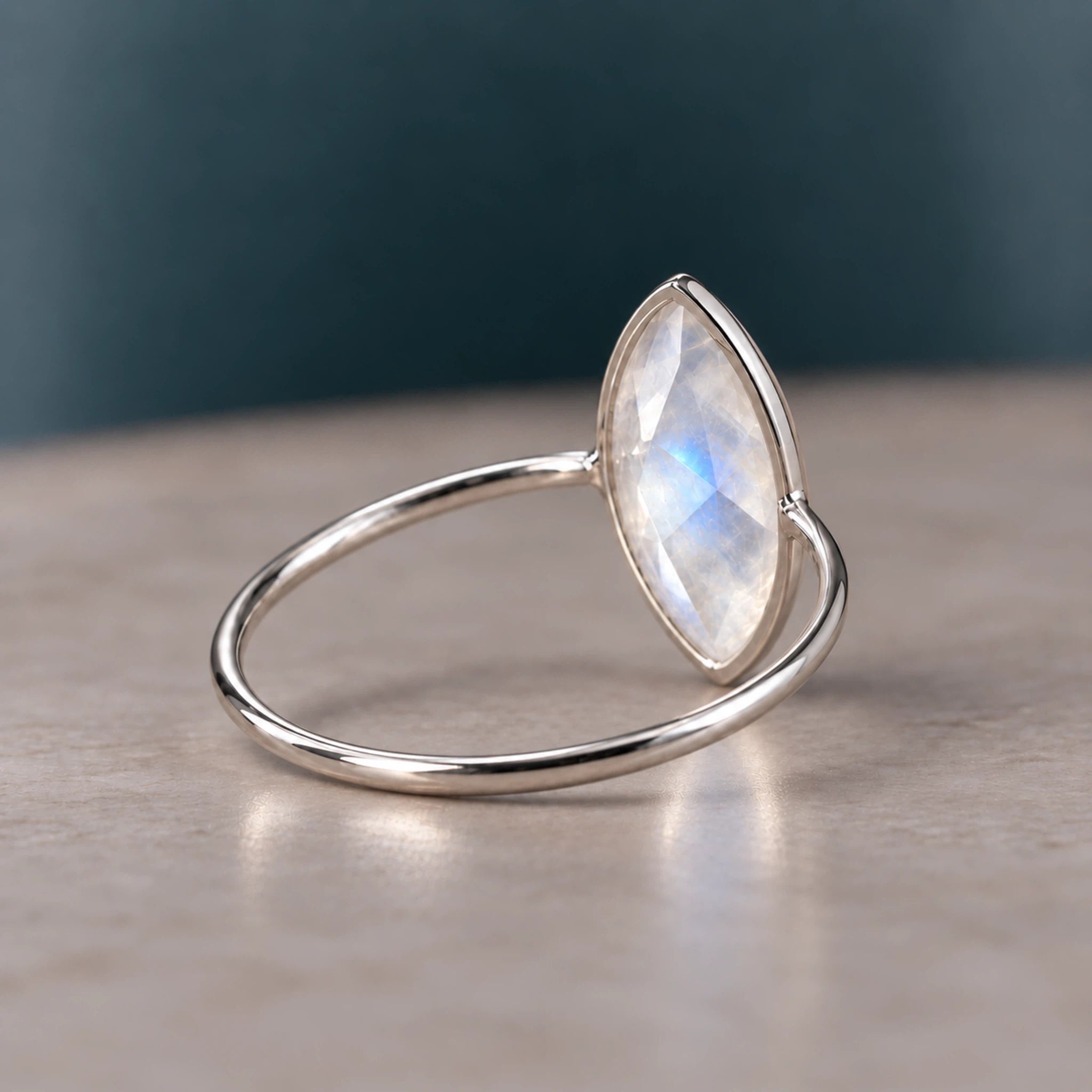 A silver ring featuring a marquise-cut iridescent moonstone gemstone, resting on a neutral beige surface with a dark teal background.