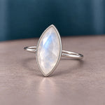 A silver ring featuring a marquise-cut iridescent moonstone gemstone, resting on a textured beige surface with a dark teal background.