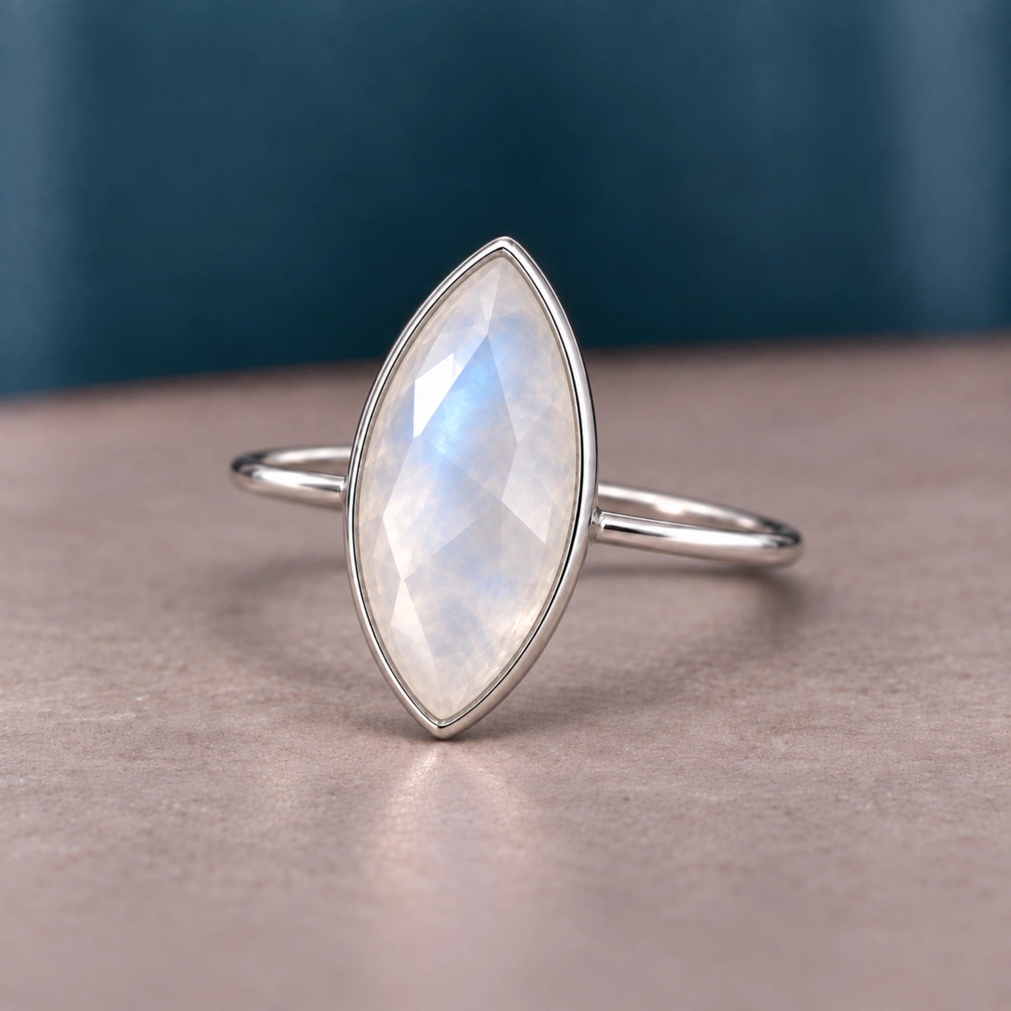 A silver ring featuring a marquise-cut iridescent moonstone gemstone, resting on a textured beige surface with a dark teal background.