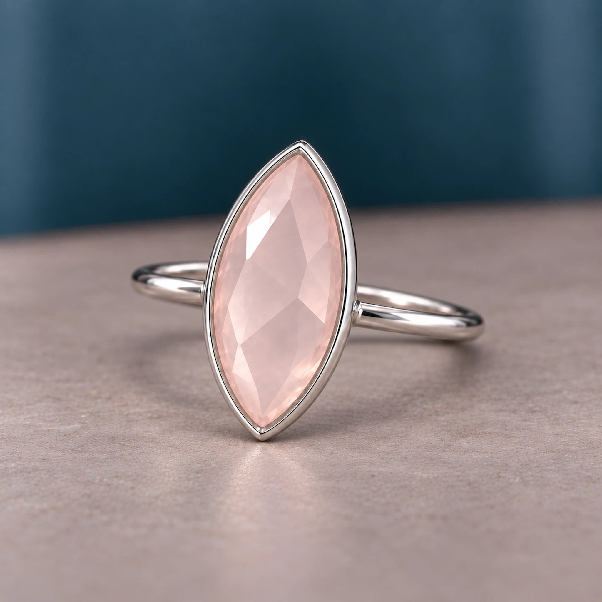 A silver ring featuring a marquise-cut pale pink rose quartz gemstone, set against a neutral gray surface with a dark teal background.