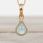 Gold pendant with a light blue aquamarine teardrop-shaped gemstone on a neutral background