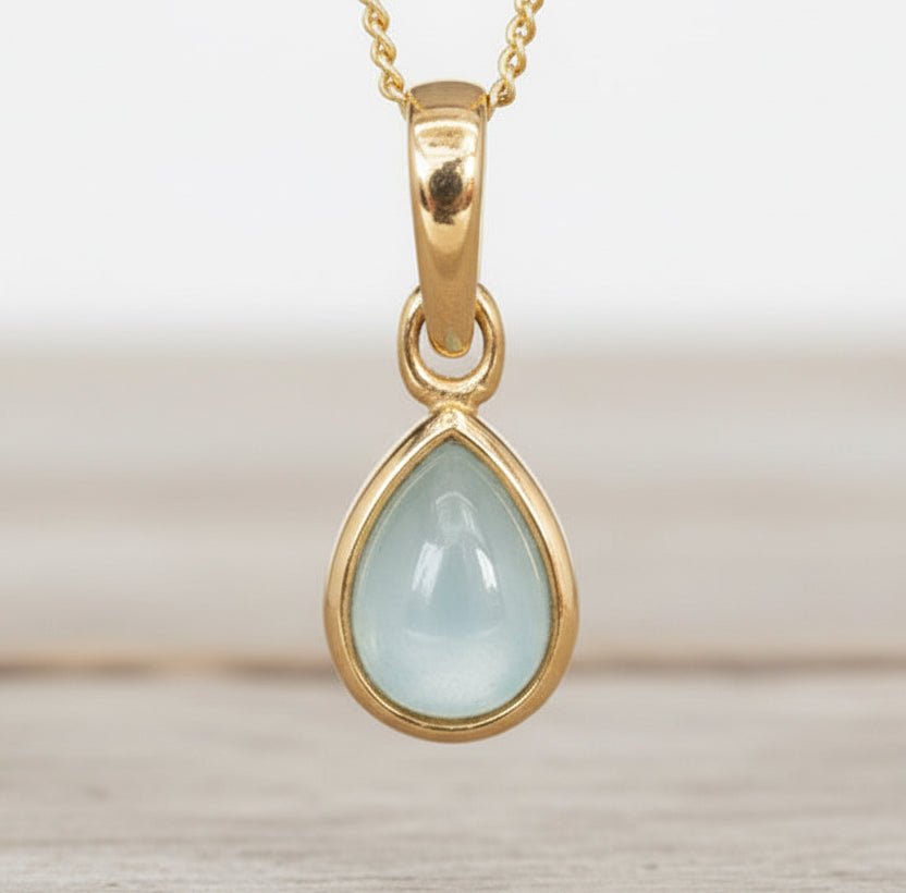 Gold pendant with a light blue aquamarine teardrop-shaped gemstone on a neutral background