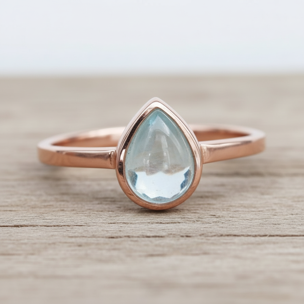 Rose gold ring with a teardrop-shaped light blue aquamarine gemstone on a wooden surface