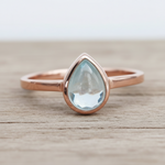 Rose gold ring with a teardrop-shaped light blue aquamarine gemstone on a wooden surface