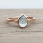 Rose gold ring with a teardrop-shaped light blue natural aquamarine gemstone on a wooden surface.